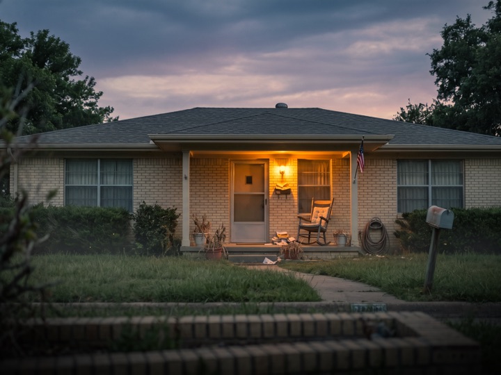 Modest inherited brick home at dusk with the porch light on and a rocking chair on the porch